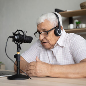 Focused mature elderly retired man in eyeglasses wearing wireless headphones looking at computer screen, talking in professional stand microphone, voice acting or recording vlog alone at home.